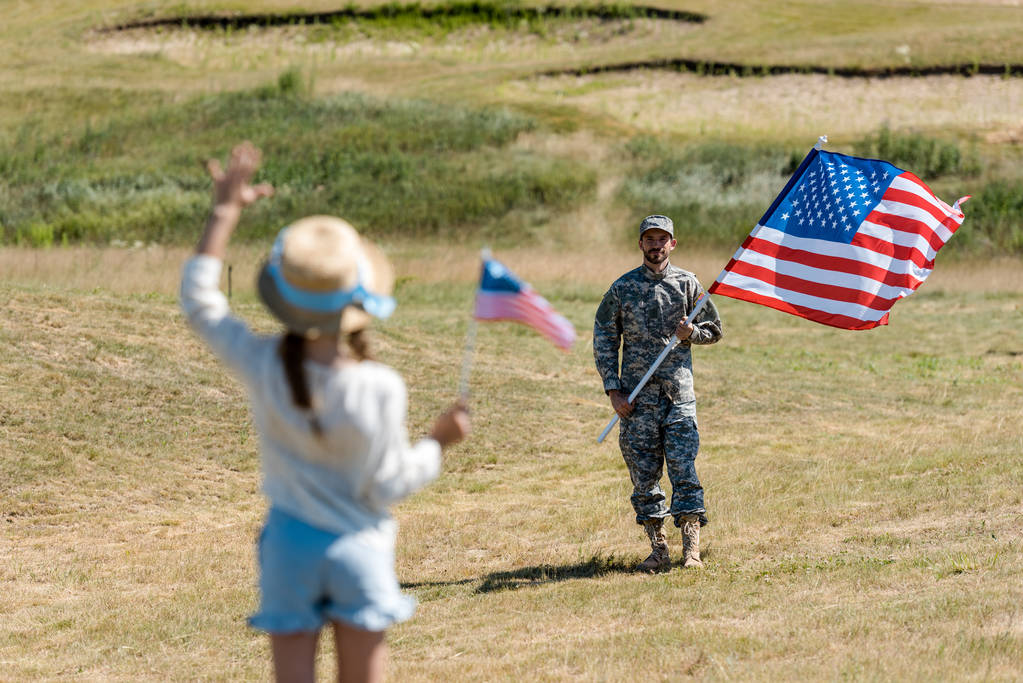 takaisin näkymä lapsi olki hattu heiluttaen käsi pitäen Amerikan lippu lähellä mies sotilaallinen univormu
  - Valokuva, kuva