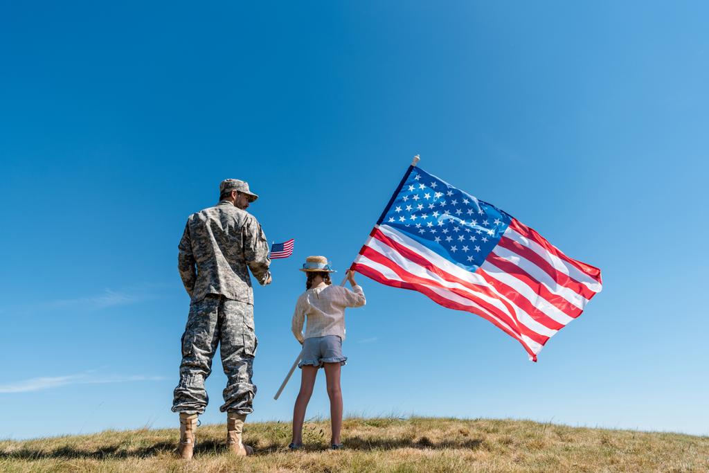 στρατιώτης με στρατιωτική στολή στέκεται κοντά σε παιδί με αμερικάνικη σημαία κατά του γαλάζιου ουρανού  - Φωτογραφία, εικόνα