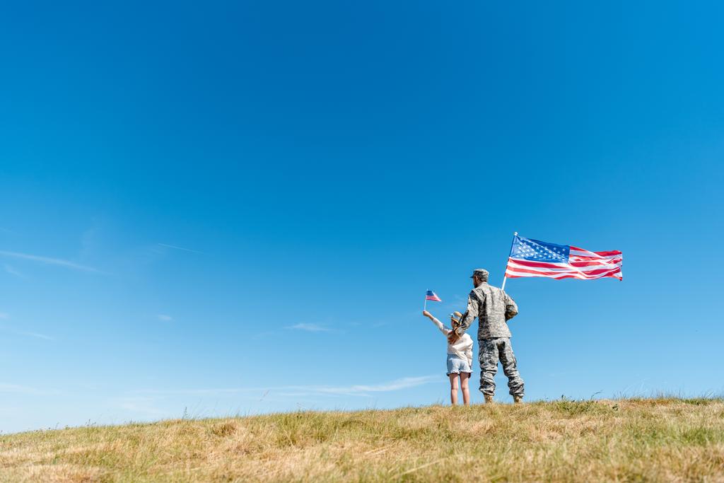 Widok z tyłu dziecka w kapeluszu słomy i wojskowych ojciec trzyma amerykańskich flag stojąc na trawie  - Zdjęcie, obraz