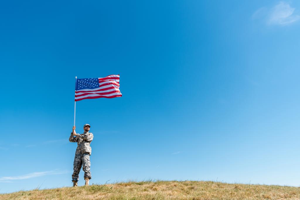 pohled s nízkým úhlem pohledu na pohledného vojenského muže v uniformách chůze s americkou vlajkou s hvězdami a pruhy  - Fotografie, Obrázek