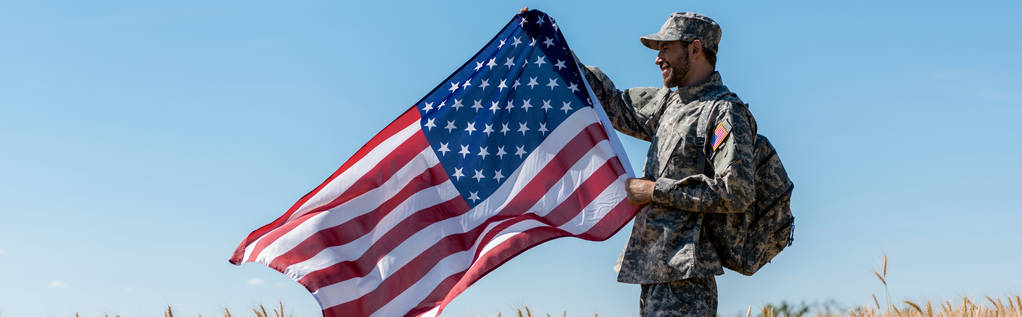 panoramische shot van knappe militaire man met rugzak houden Amerikaanse vlag  - Foto, afbeelding