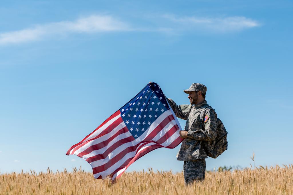 szczęśliwy żołnierz w mundurze wojskowym stojący w polu ze złotą pszenicą i trzymając amerykańską flagę  - Zdjęcie, obraz