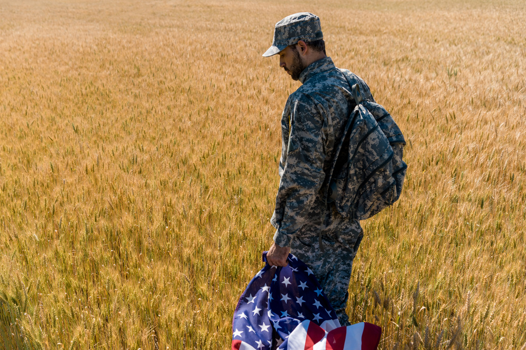 小麦と一緒に野原に立ちながらアメリカ国旗を掲げた軍服を着た愛国的な兵士  - 写真・画像