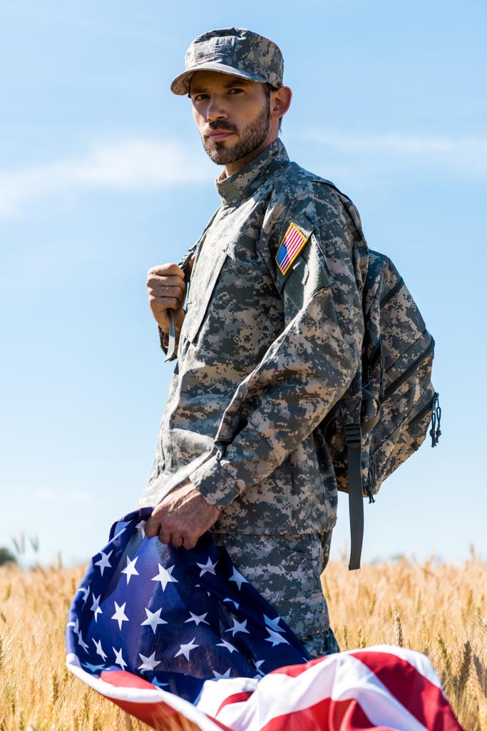 selektivní zaměření vojáka v uniformním držení americké vlajky při stání v terénu  - Fotografie, Obrázek