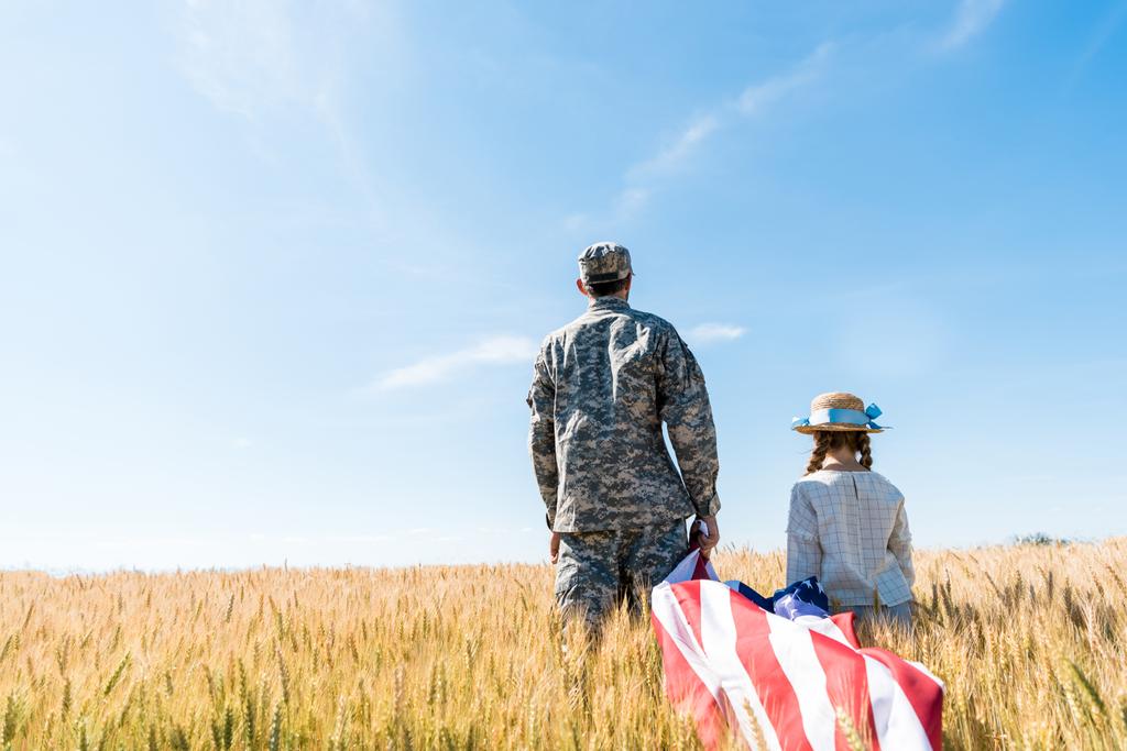 制服を着た兵士と子供が野原に立ち、アメリカ国旗を掲げる背中の眺め  - 写真・画像