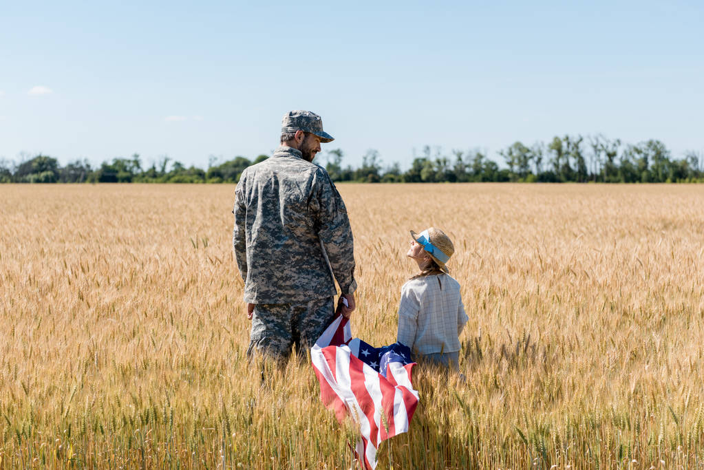 制服姿の兵士が、フィールドでアメリカ国旗を掲げながら娘を見ている  - 写真・画像