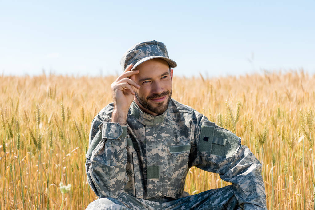 positieve militaire man aanraken GLB en glimlachend in veld  - Foto, afbeelding