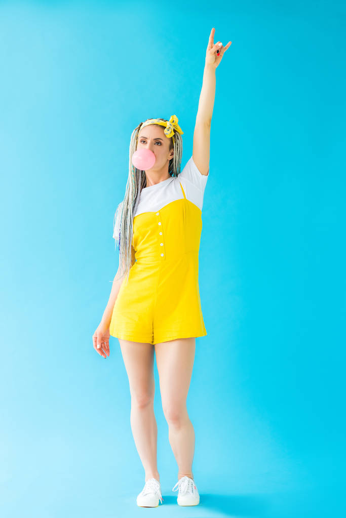 Девушка с дредами, пускающая жвачку и показывающая рок-знак на бирюзовой
 - Фото, изображение