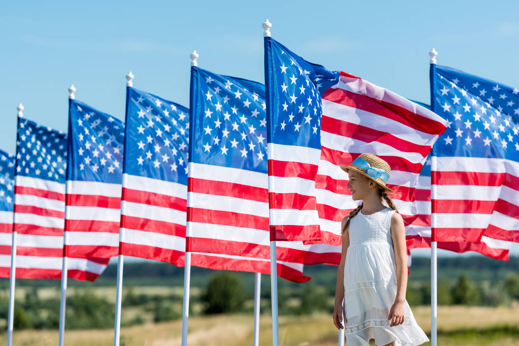 niño alegre de pie en vestido blanco y sombrero de paja cerca de banderas americanas
  - Foto, Imagen