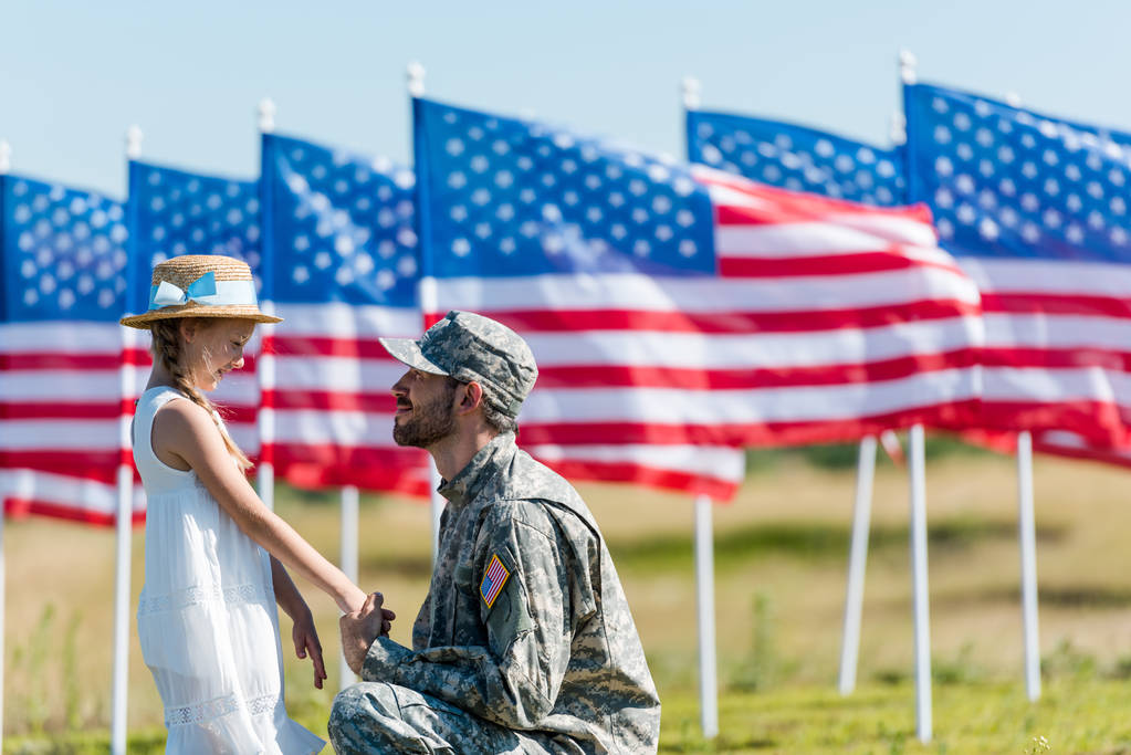 gelukkige militaire man kijken naar Kid in stro hoed terwijl het houden van handen in de buurt van Amerikaanse vlaggen met sterren en strepen  - Foto, afbeelding