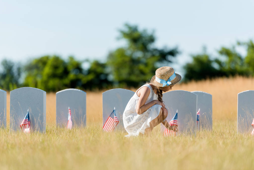 enfoque selectivo de niño en vestido y sombrero de paja sentado cerca de lápida con bandera americana en el cementerio
  - Foto, Imagen