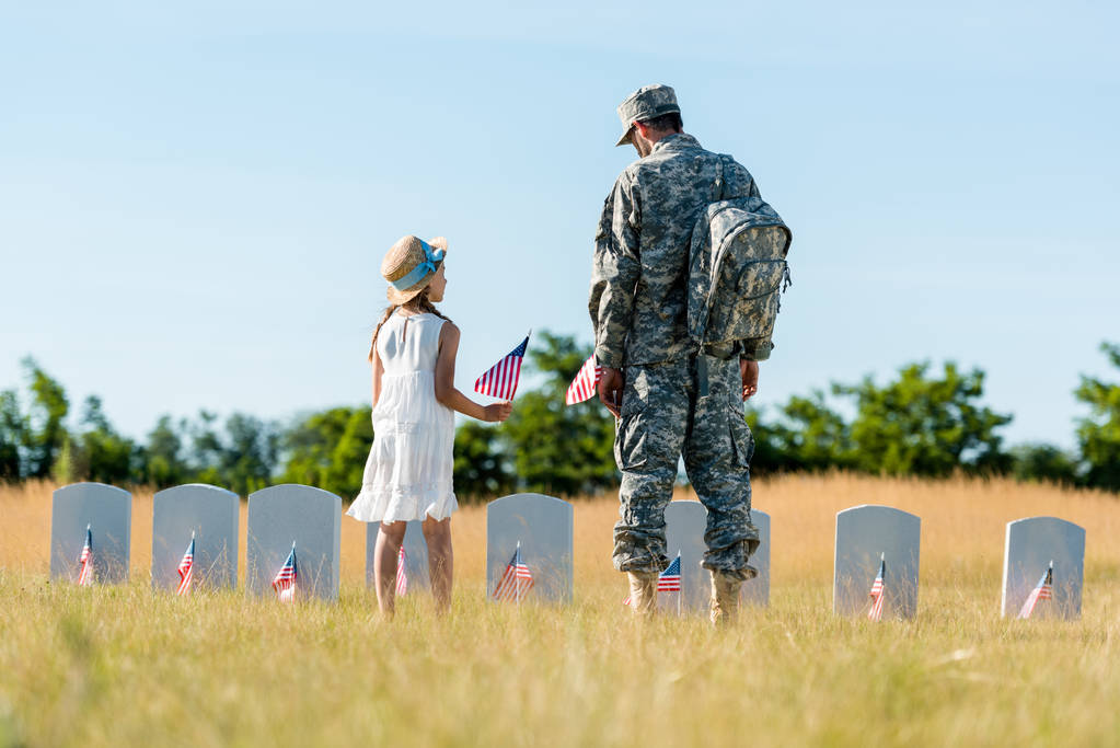 παιδί με ψάθινο καπέλο και άνθρωπος με στρατιωτική στολή στέκεται κοντά σε κεφαλαίους και κρατώντας αμερικανικές σημαίες στο νεκροταφείο  - Φωτογραφία, εικόνα