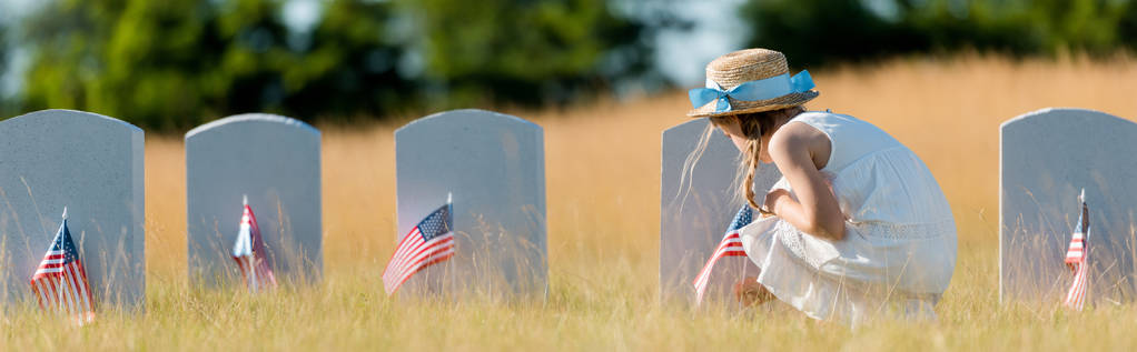 墓地にアメリカの旗を持つヘッドストーンの近くに座っているドレスと麦わら帽子の子供のパノラマショット  - 写真・画像
