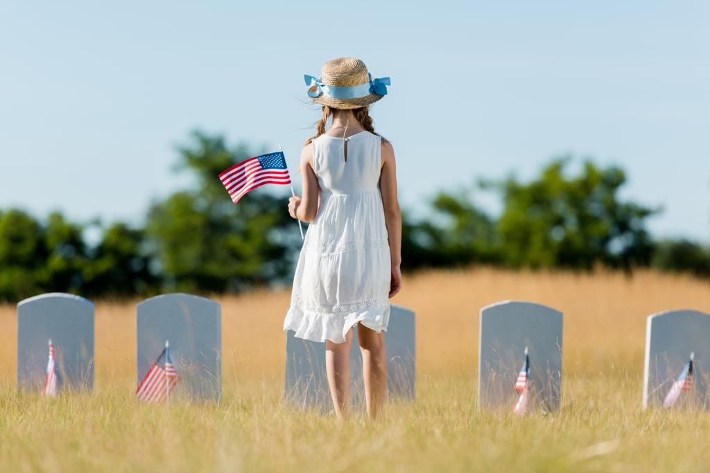 pohled zpět na dítě v šatech a slaměném klobouku stojících blízko náhrobku a držící americkou vlajku na hřbitově  - Fotografie, Obrázek