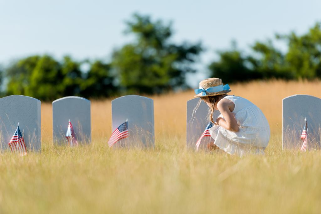Selektiver Fokus des Kindes in Kleid und Strohhut sitzt neben Grabstein mit amerikanischer Flagge auf Friedhof  - Foto, Bild