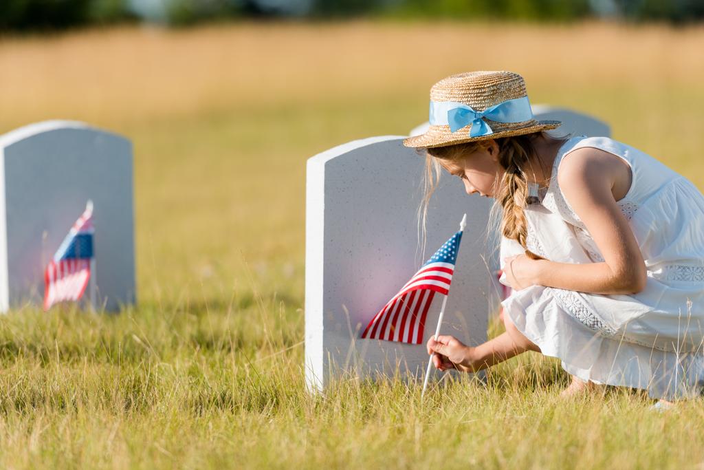 selective focus of adorable kid in straw hat sitting near headstone with american flag in graveyard  - Photo, Image