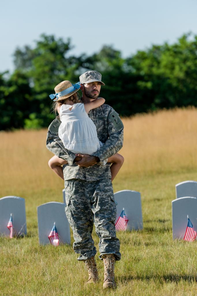військовий чоловік у капелюсі, що тримає дитину в солом'яному капелюсі біля надгробків з американськими прапорами
  - Фото, зображення