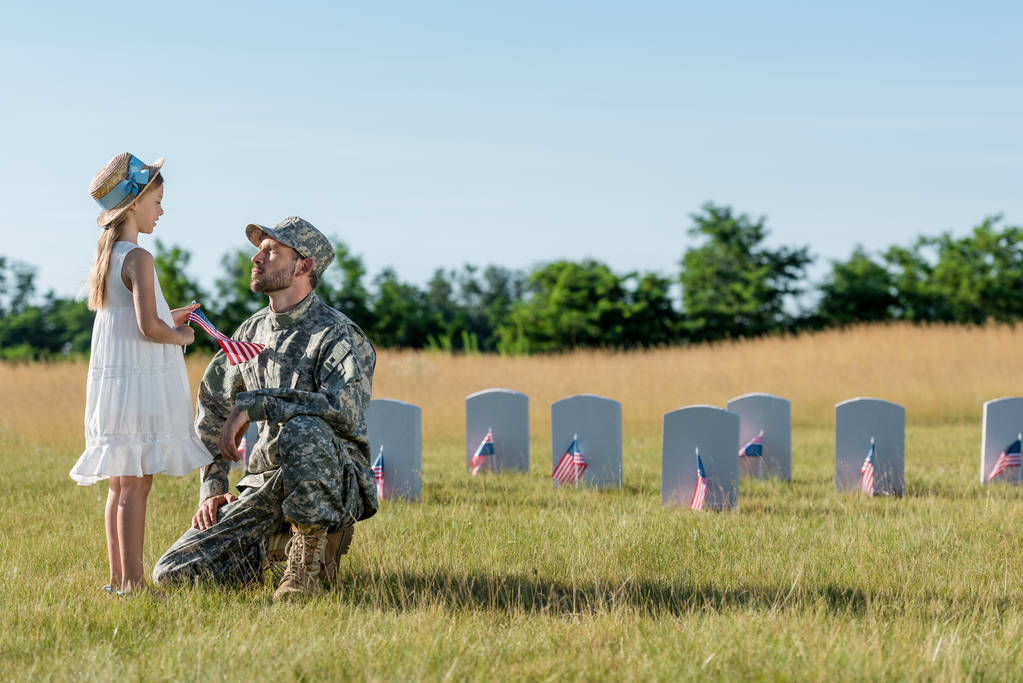 sotilas lippis istuu lapsen lähellä olki hattu ja hautakivet hautausmaalla
  - Valokuva, kuva