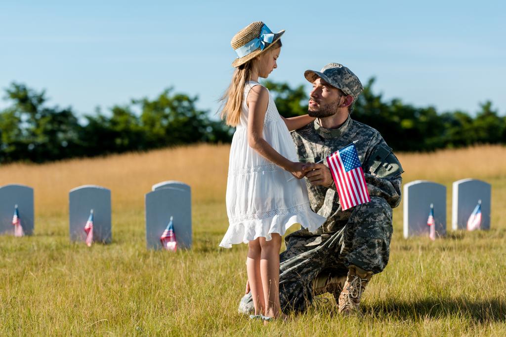 katonai ember a sapka mellett ül a gyermek és a sírkövek a temetőben  - Fotó, kép