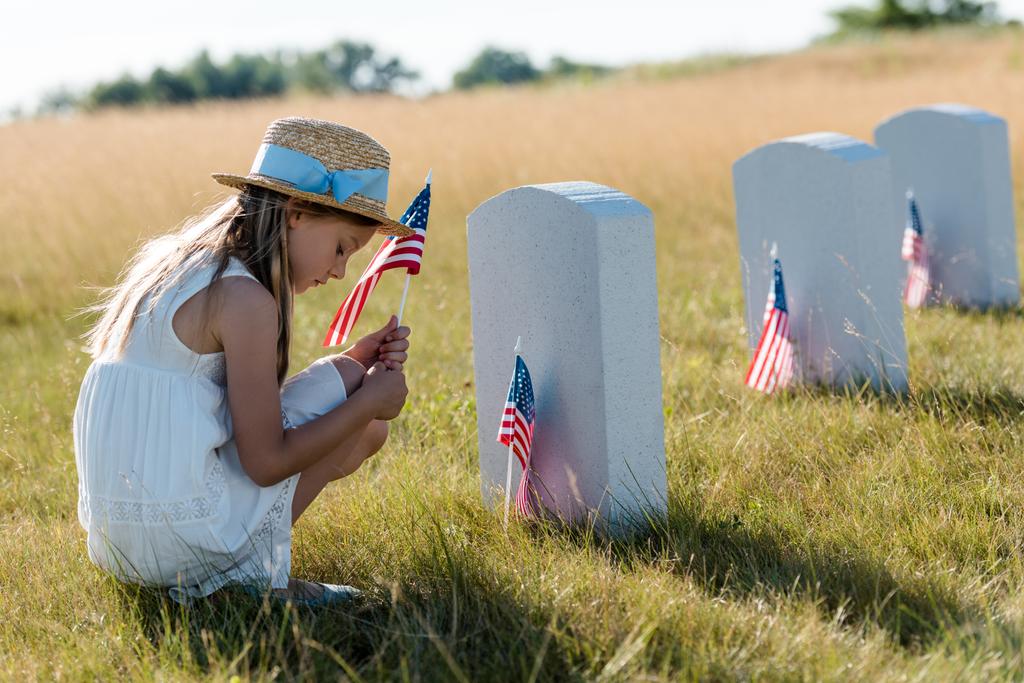 sad kid in straw hat sitting near headstones and holding american flag  - Photo, Image