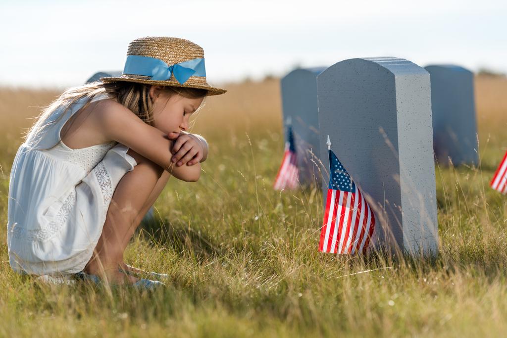 sad kid in straw hat sitting near headstones with american flags  - Photo, Image