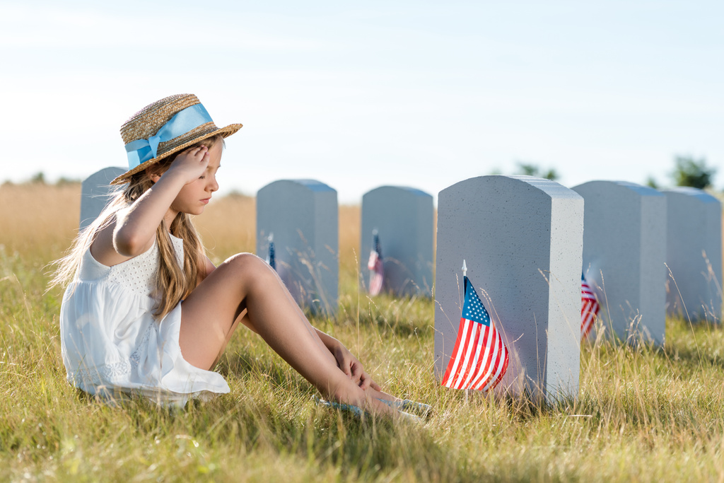 bambino in cappello di paglia che saluta mentre seduto vicino a lapidi con bandiere americane
  - Foto, immagini