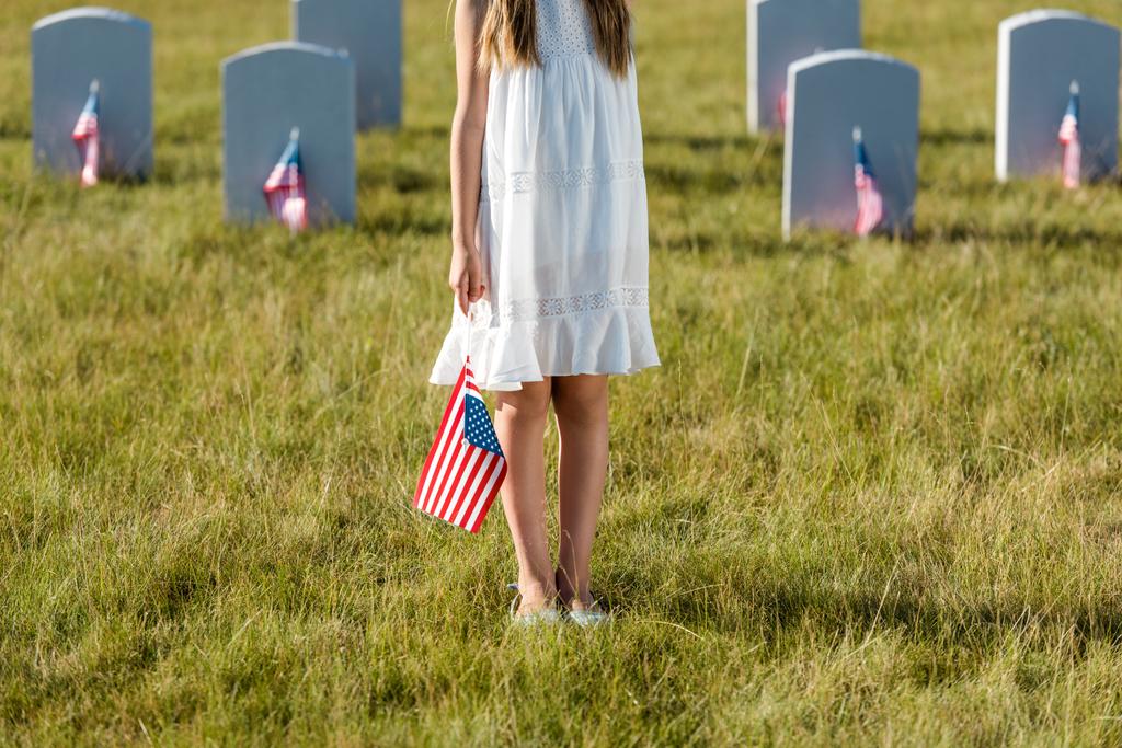 περικομμένη θέα του παιδιού σε λευκό φόρεμα στέκεται σε νεκροταφείο με αμερικανική σημαία  - Φωτογραφία, εικόνα