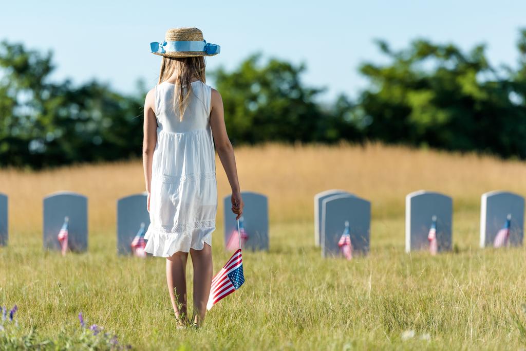 вид ззаду дитини в білій сукні, що стоїть на кладовищі з американським прапором
  - Фото, зображення