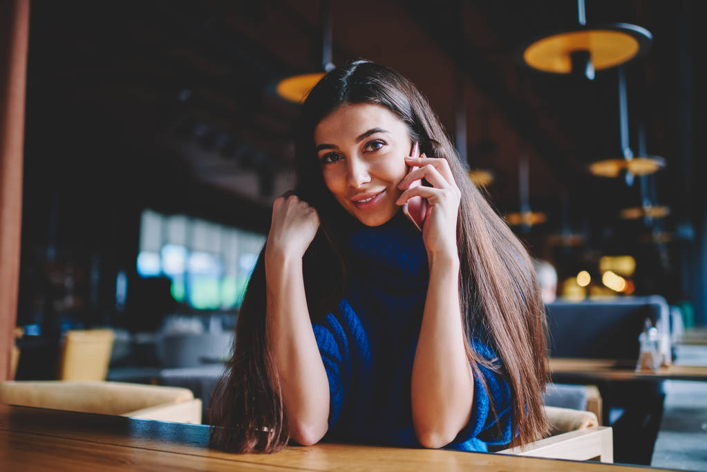 Portret wesołej dziewczyny Hipster odpoczynku w kawiarni podczas komunikacji przez smartfona, uśmiechnięta pozytywna kobieta patrząc na kamerę i podejmowania rozmowy przez telefon komórkowy podłączony do WiFi w pomieszczeniu - Zdjęcie, obraz