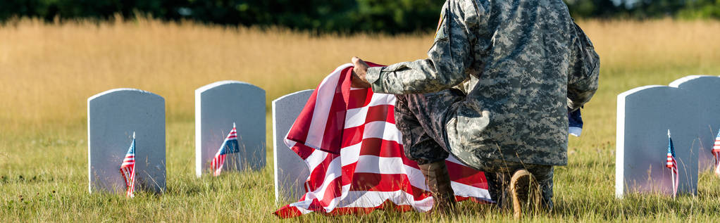 panoramic shot of man in military uniform holding american flag while sitting in graveyard  - Photo, Image