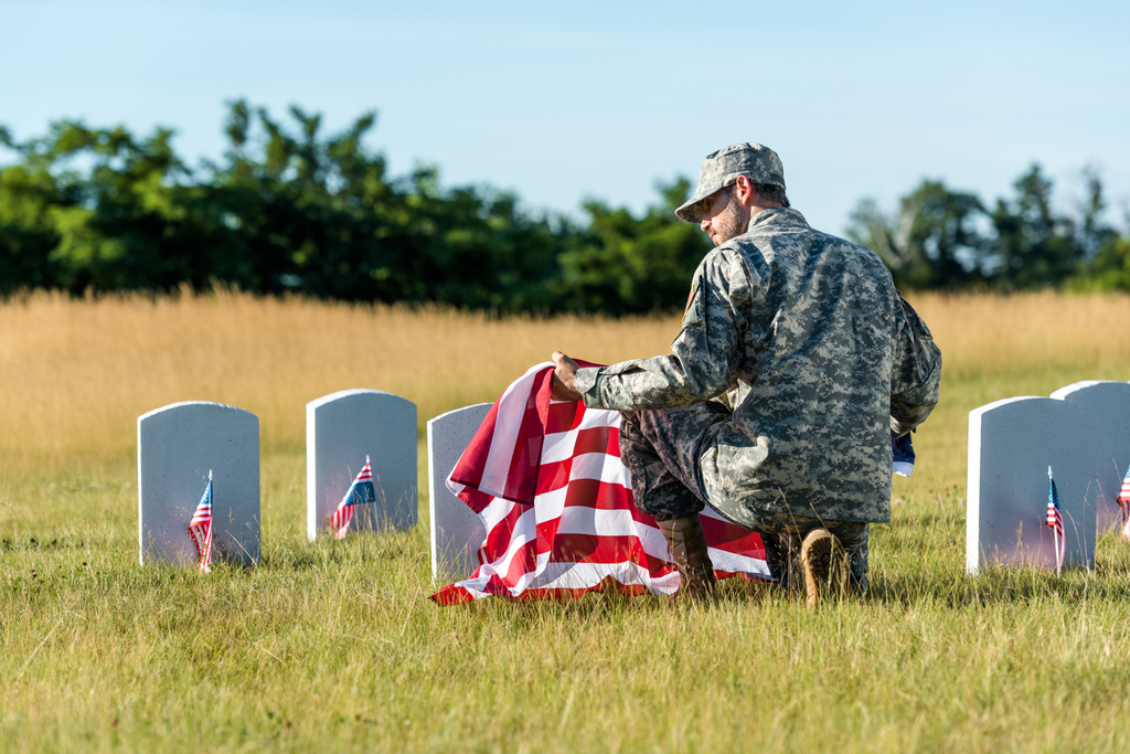άνθρωπος με στρατιωτική στολή και καπάκι που κρατά αμερικάνικη σημαία ενώ κάθεται στο νεκροταφείο  - Φωτογραφία, εικόνα