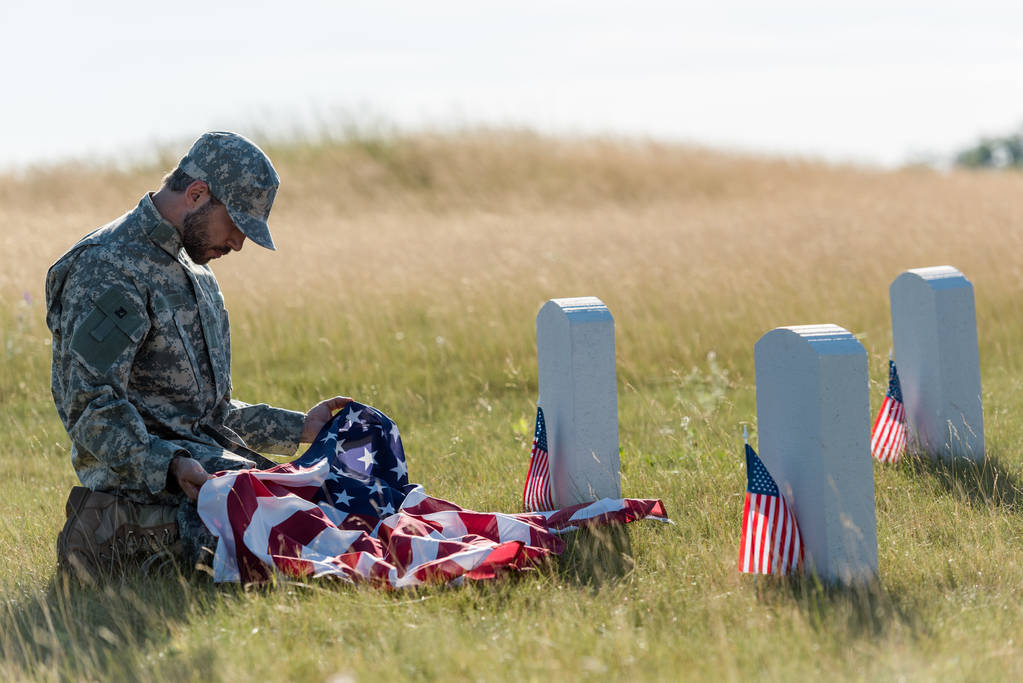 λυπημένος στρατιώτης σε καμουφλάζ στολή και καπάκι κρατώντας αμερικάνικη σημαία ενώ κάθονται στο νεκροταφείο  - Φωτογραφία, εικόνα