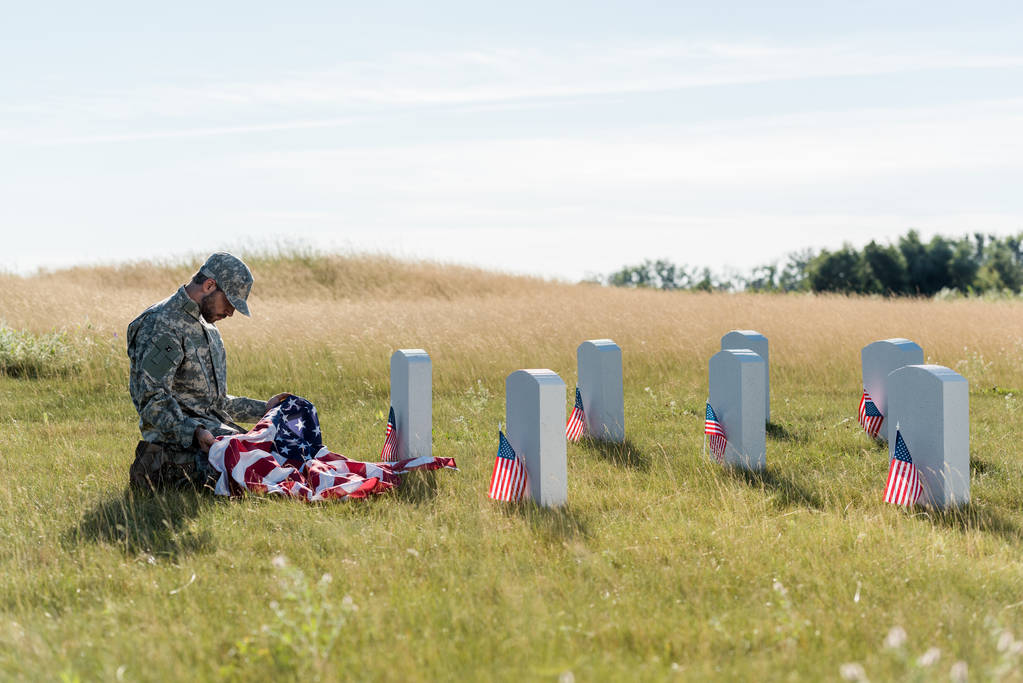 сумний солдат у камуфляжній формі і кепка тримає американський прапор, сидячи на кладовищі
  - Фото, зображення