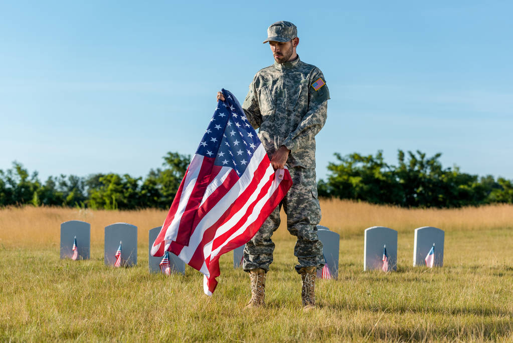 przystojny weteran w kamuflażu jednolitego posiadania amerykańskiej flagi i stoi na cmentarzu  - Zdjęcie, obraz