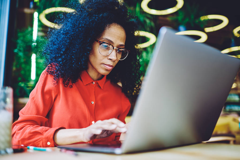 Σοβαρή Αφρο-Αμερικάνικη γυναίκα που εργάζεται διαδικτυακά με πληροφορίες δεδομένων στο αρχείο κειμένου στη σύγχρονη συσκευή φορητού υπολογιστή χρησιμοποιώντας Wi-Fi σύνδεση στο Internet στο κυλικείο, εξειδικευμένο μαύρο κορίτσι ανταλλαγής μηνυμάτων με τους συναδέλφους - Φωτογραφία, εικόνα