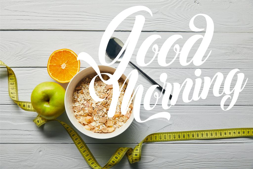 верхний вид измерительной ленты, ложки и завтрака хлопья в миске рядом яблоко и апельсин на деревянном белом фоне с добрым утром надписи
 - Фото, изображение