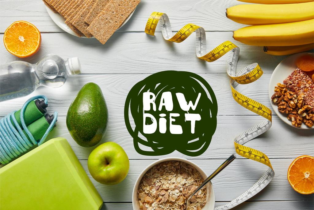 Top zicht op dieet voedsel in de buurt van meetlint en sportuitrusting op houten witte achtergrond met RAW Diet belettering - Foto, afbeelding