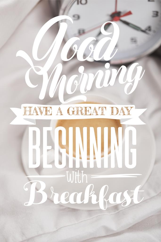 Blick von oben auf Kaffee in weißer Tasse auf Untertasse in der Nähe von silbernem Wecker auf Bettwäsche mit Guten Morgen, haben einen tollen Tag beginnen mit Frühstück Schriftzug - Foto, Bild
