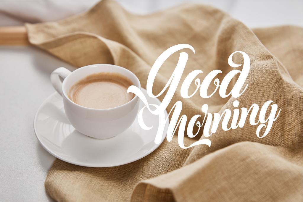 おはようイラストとベージュナプキンの近くのソーサーに白いカップでコーヒーのクローズアップビュー - 写真・画像