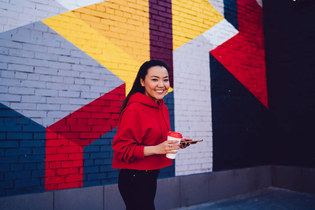 Halblanges Porträt einer fröhlichen Bloggerin mit Handy im Freien, die in die Kamera schaut und lächelt, positives Hipstermädchen, das Zell- und Koffeingetränk in der Nähe einer bunten Wand mit Kopierraum hält - Foto, Bild