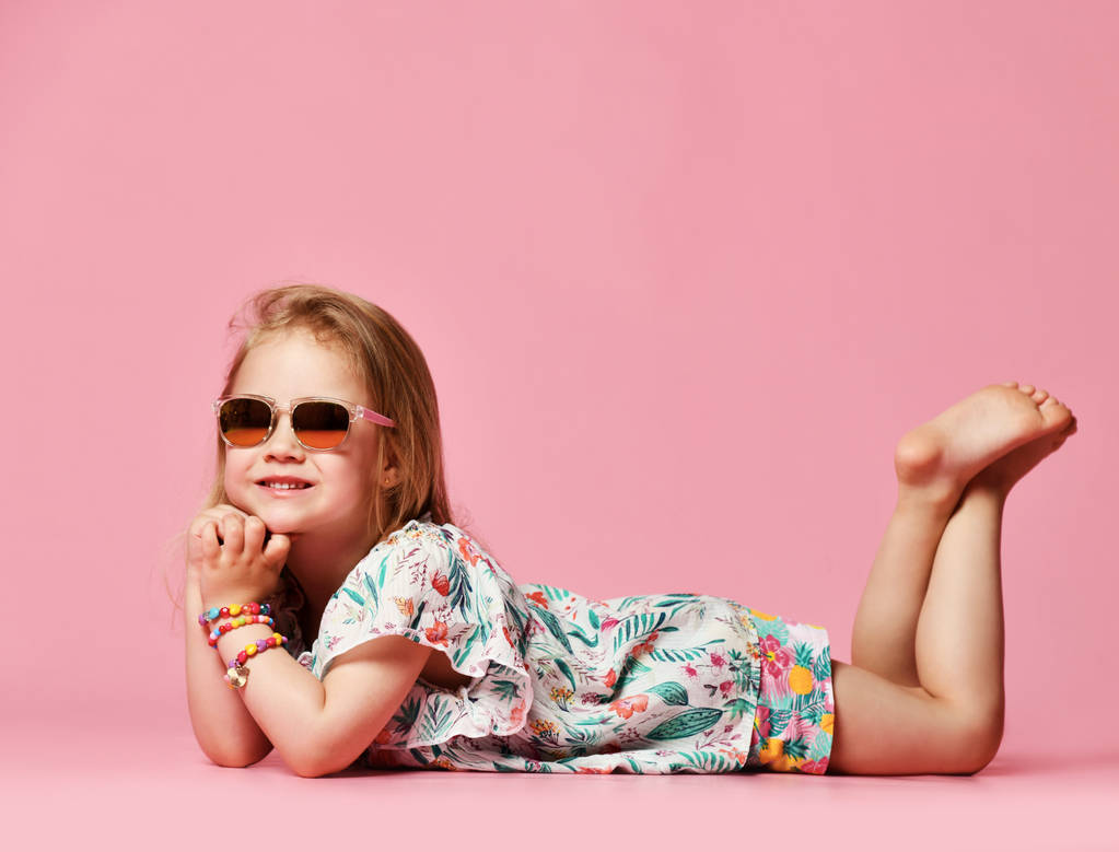 小さな女の子はピンクの背景のサングラスで夏服でスタジオの床に横たわっている側を見ます - 写真・画像