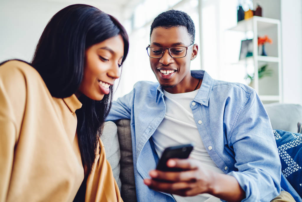 Χαμογελώντας African American ζευγάρι στην αγάπη βλέποντας βίντεο σε απευθείας σύνδεση στο smartphone ξοδεύοντας ελεύθερο χρόνο στο σπίτι, χαρούμενα σκούρο δέρμα hipster τύπος δείχνει φωτογραφίες δώρων στο κινητό τηλέφωνο στην όμορφη φίλη του - Φωτογραφία, εικόνα