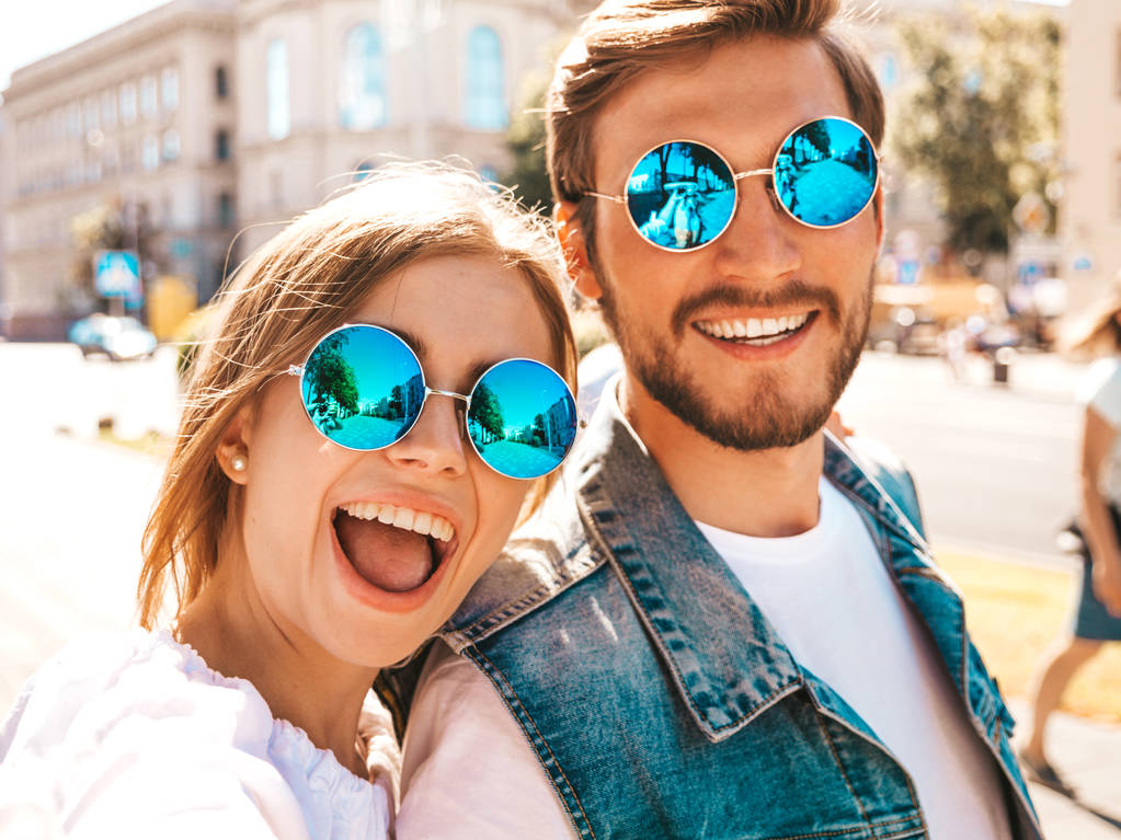 Uśmiechnięta piękna dziewczyna i jej przystojny chłopak w luźnych letnich ubrań.Szczęśliwa rodzina biorąc selfie autoportret siebie na smartfonie aparat w sunglasses.having zabawy na tle ulicy  - Zdjęcie, obraz