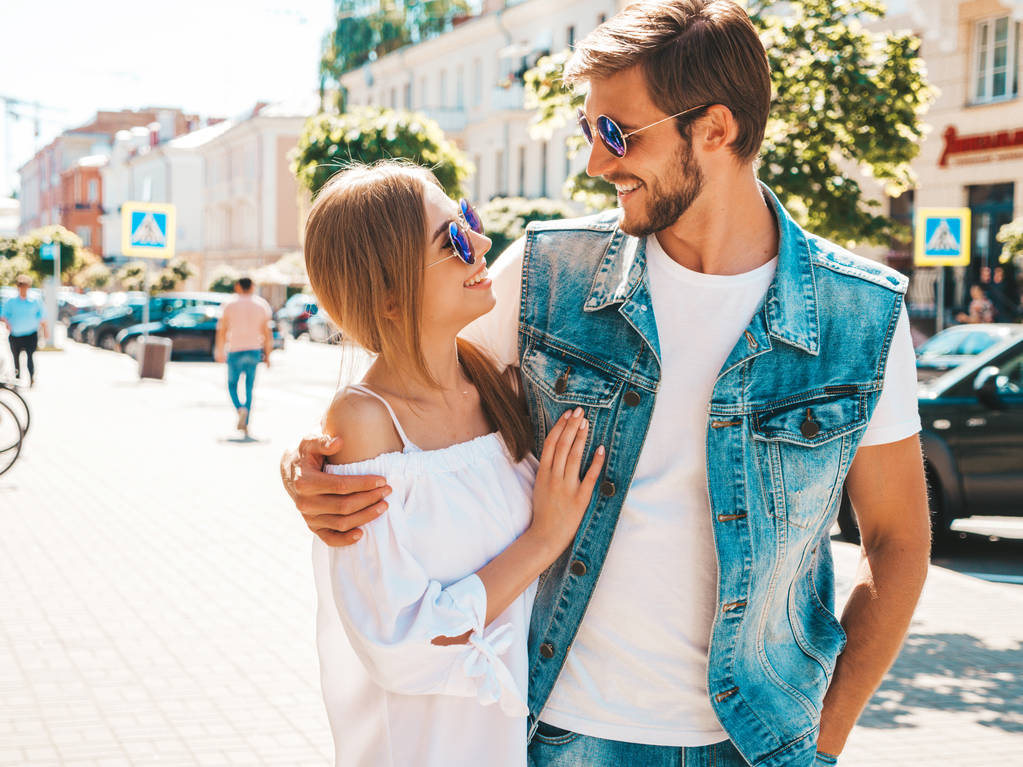 Gülümseyen güzel kız ve onun yakışıklı erkek arkadaşı sokakta yürüyor. Rahat yazlık elbiseli kadın ve kot pantolon giyen bir erkek. Mutlu neşeli çift aile güneş gözlüğü eğlenmek - Fotoğraf, Görsel
