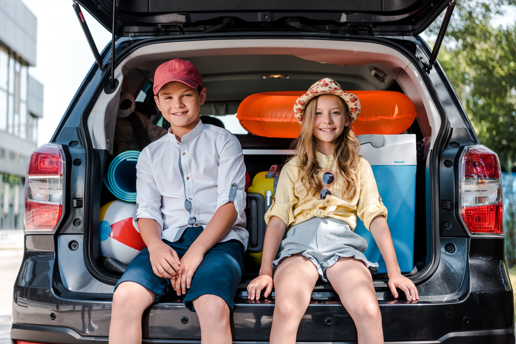 mutlu kız ve erkek kardeşi araba bagajında oturan ve kamera bakarak - Fotoğraf, Görsel