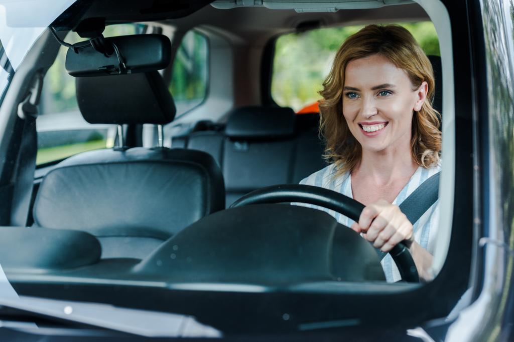 επιλεκτική εστίαση της θετικής γυναίκας που κρατά το τιμόνι και το αυτοκίνητο οδήγησης  - Φωτογραφία, εικόνα