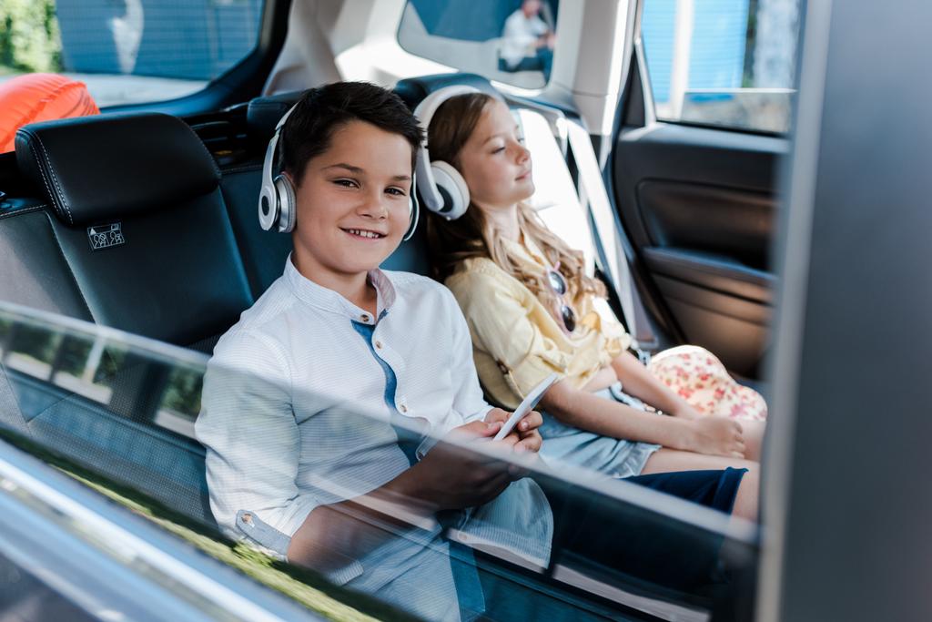 車の中で姉妹の近くのヘッドフォンでスマートフォンを使用し、音楽を聴く少年の選択的な焦点  - 写真・画像