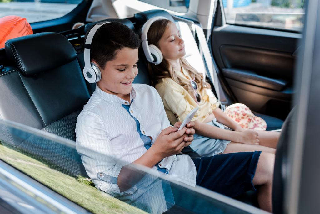 foco seletivo do menino usando smartphone e ouvir música em fones de ouvido perto da irmã com olhos fechados no carro
  - Foto, Imagem