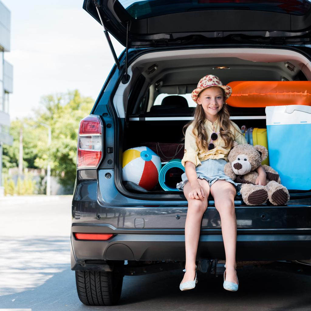 happy kid in straw hat  holding teddy bear and sitting in car trunk near building  - Photo, Image