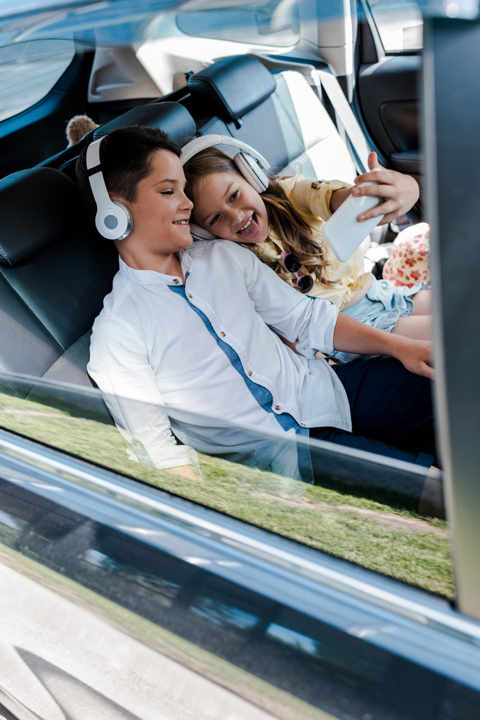 Selektiver Fokus eines fröhlichen Kindes mit Kopfhörern, das Selfie mit Bruder im Auto macht  - Foto, Bild